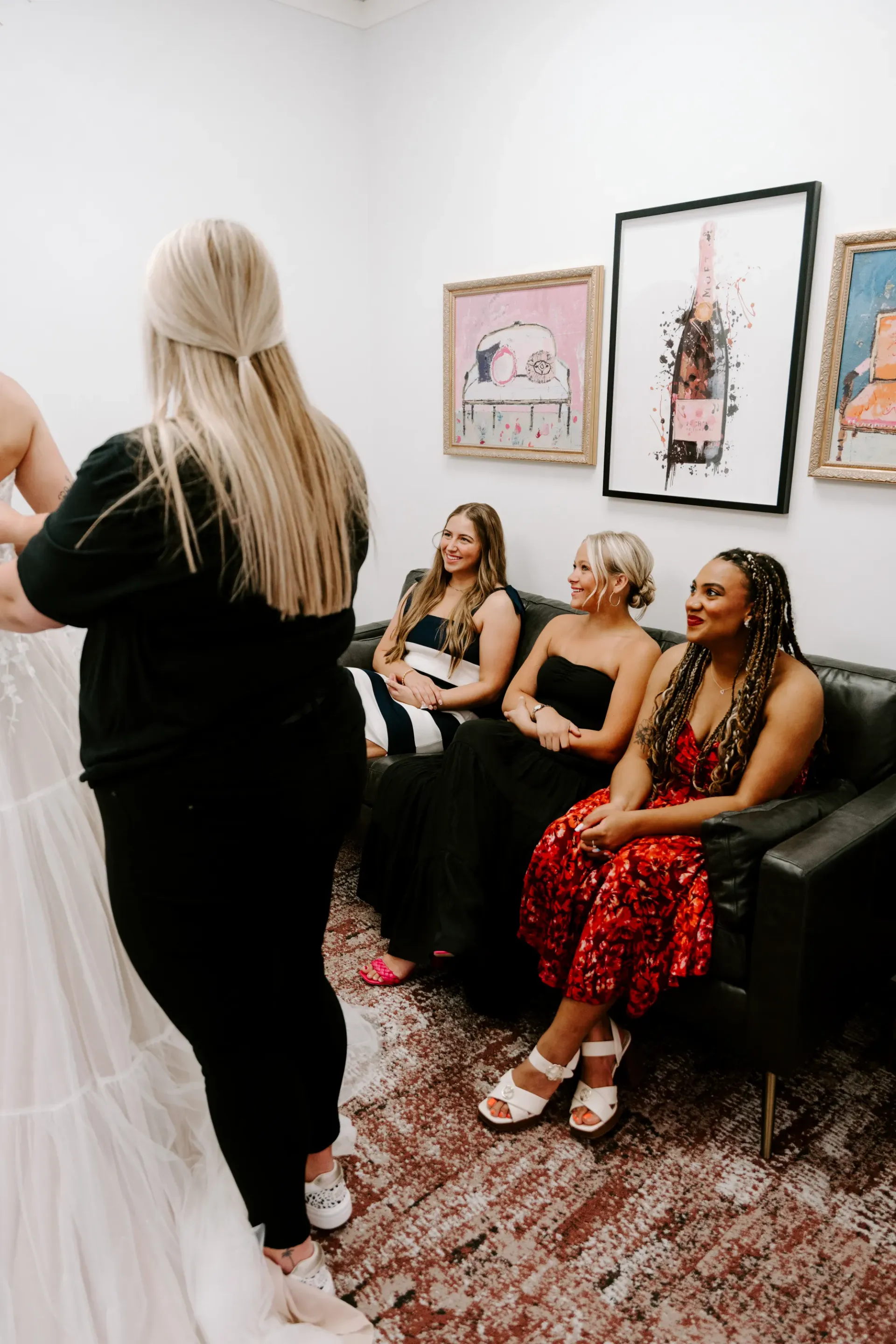 Woman trying on a dress; three friends on a black sofa watch. White room with art.