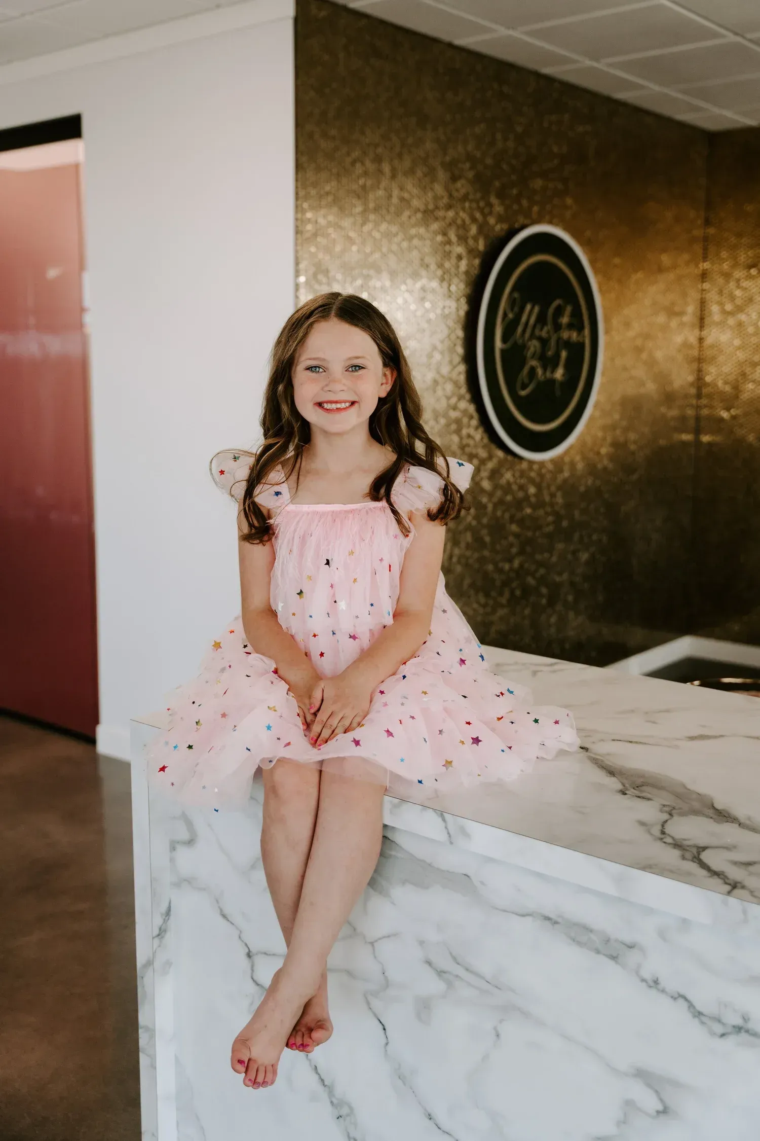 Girl in pink dress with red polka dots sits on marble counter, smiling.