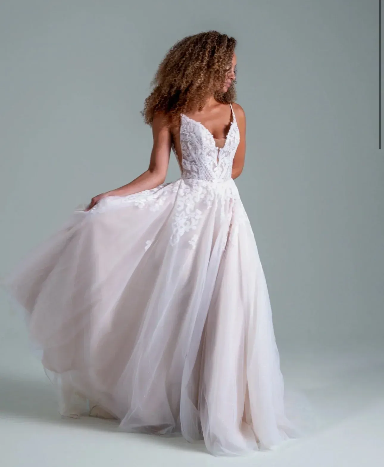 Woman in a white, flowing wedding dress, light background. She twirls, smiling, with curly hair.