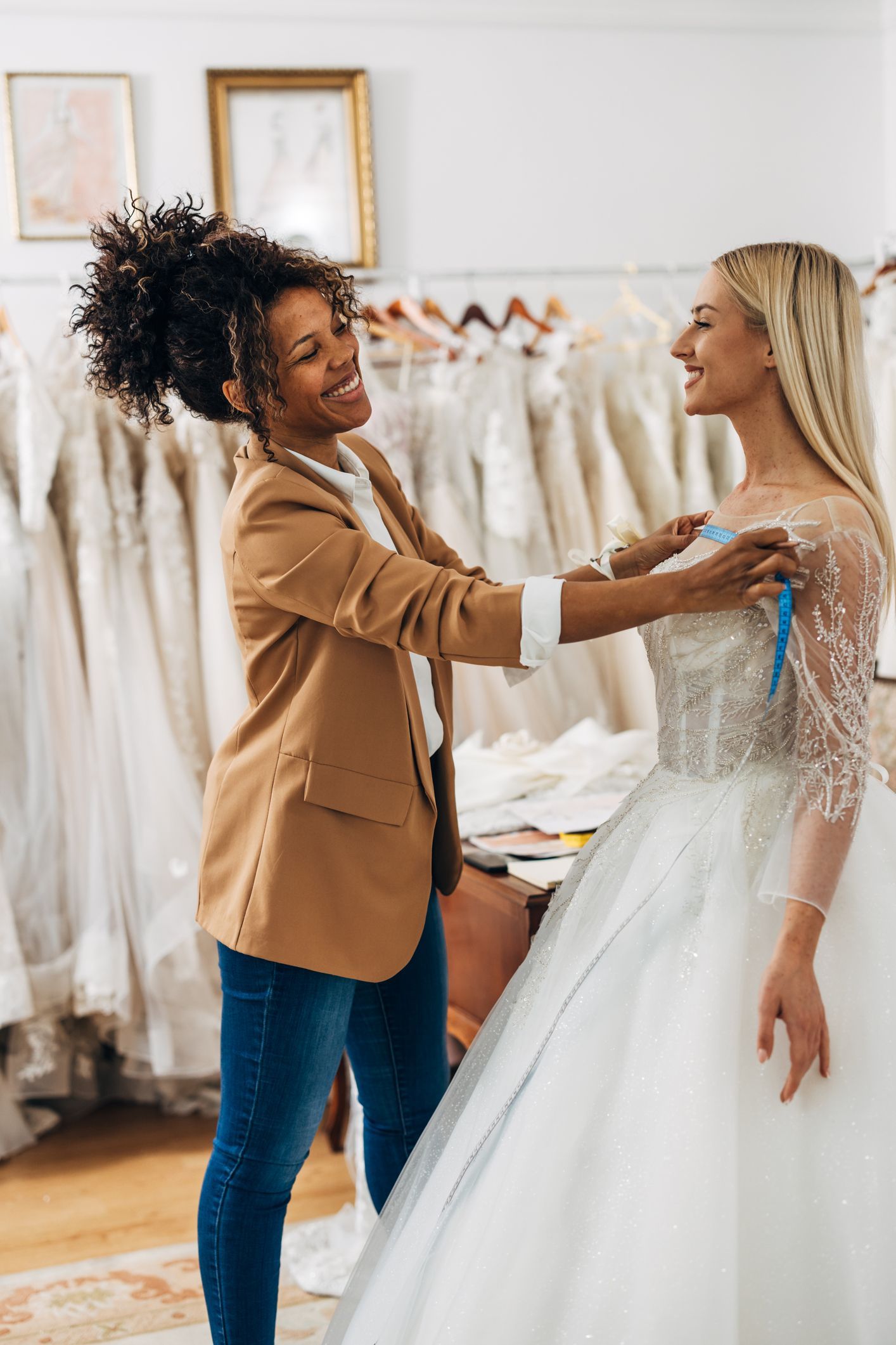 Woman in brown blazer measures a bride in a wedding dress in a bridal shop. Both women are smiling.