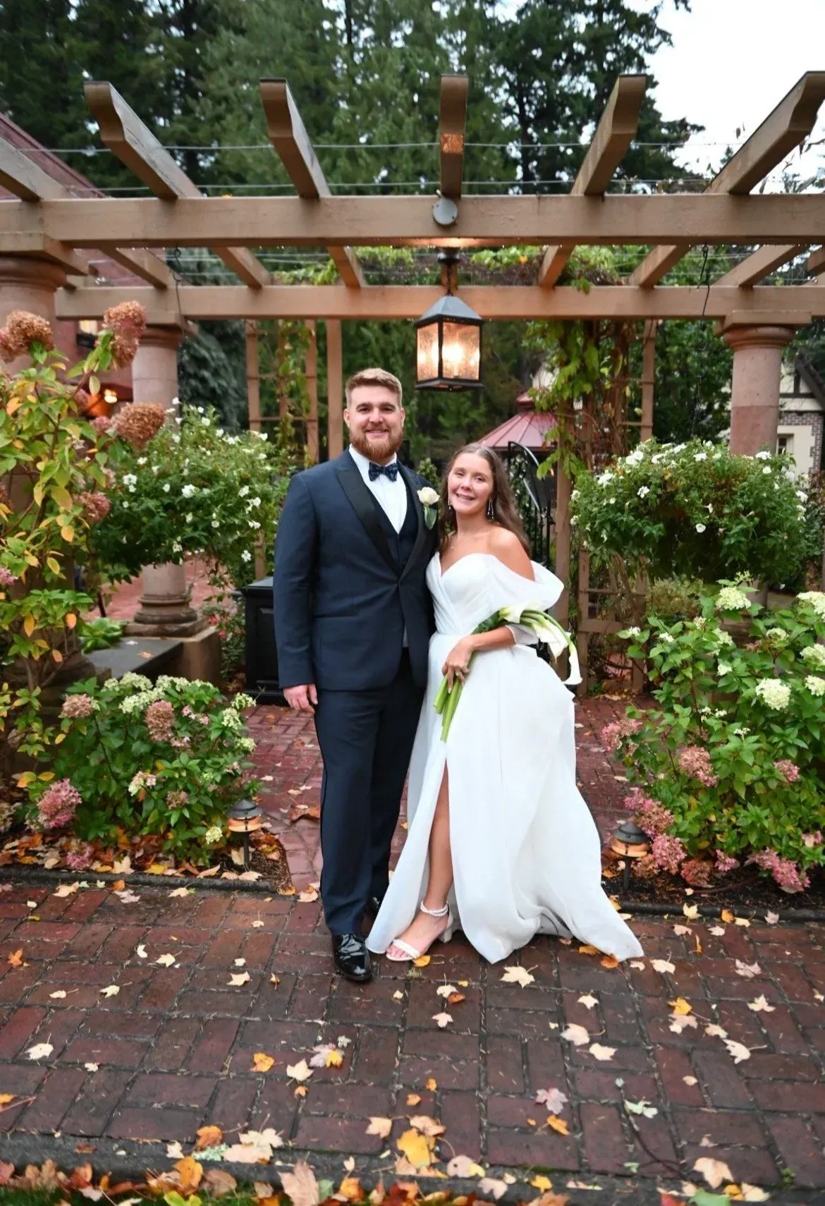 Bride and groom pose at a wedding, under a wooden arbor with flowers. The couple is smiling.