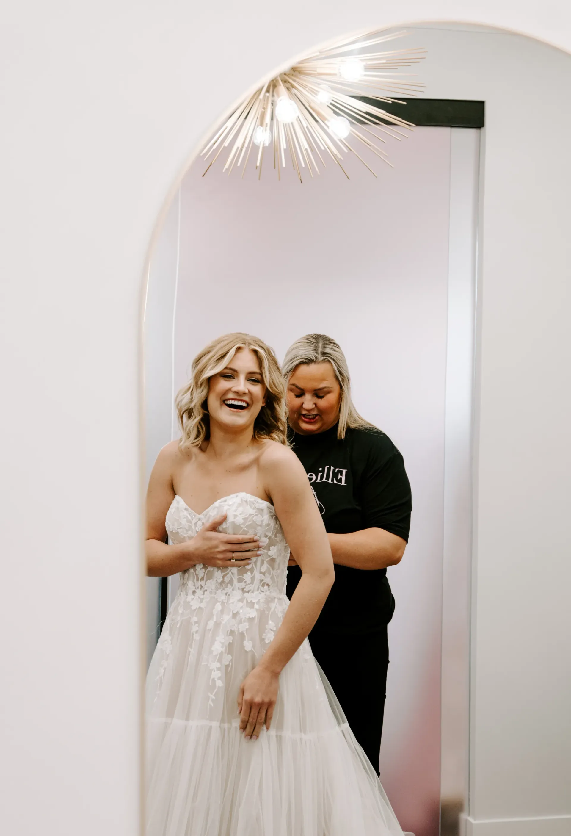 Bride smiling as attendant adjusts her wedding dress in a bridal shop.