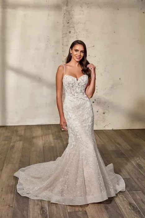 Woman in white mermaid wedding dress, smiling, hands near hair, standing in front of beige wall, wooden floor.