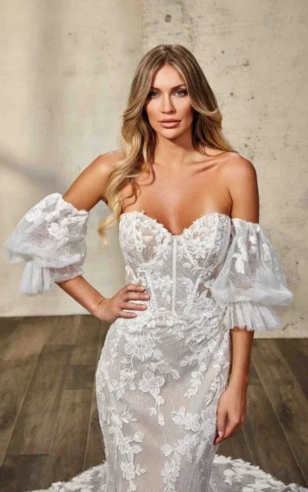 Woman in a white, lace off-the-shoulder wedding dress, posed indoors with hand on hip.