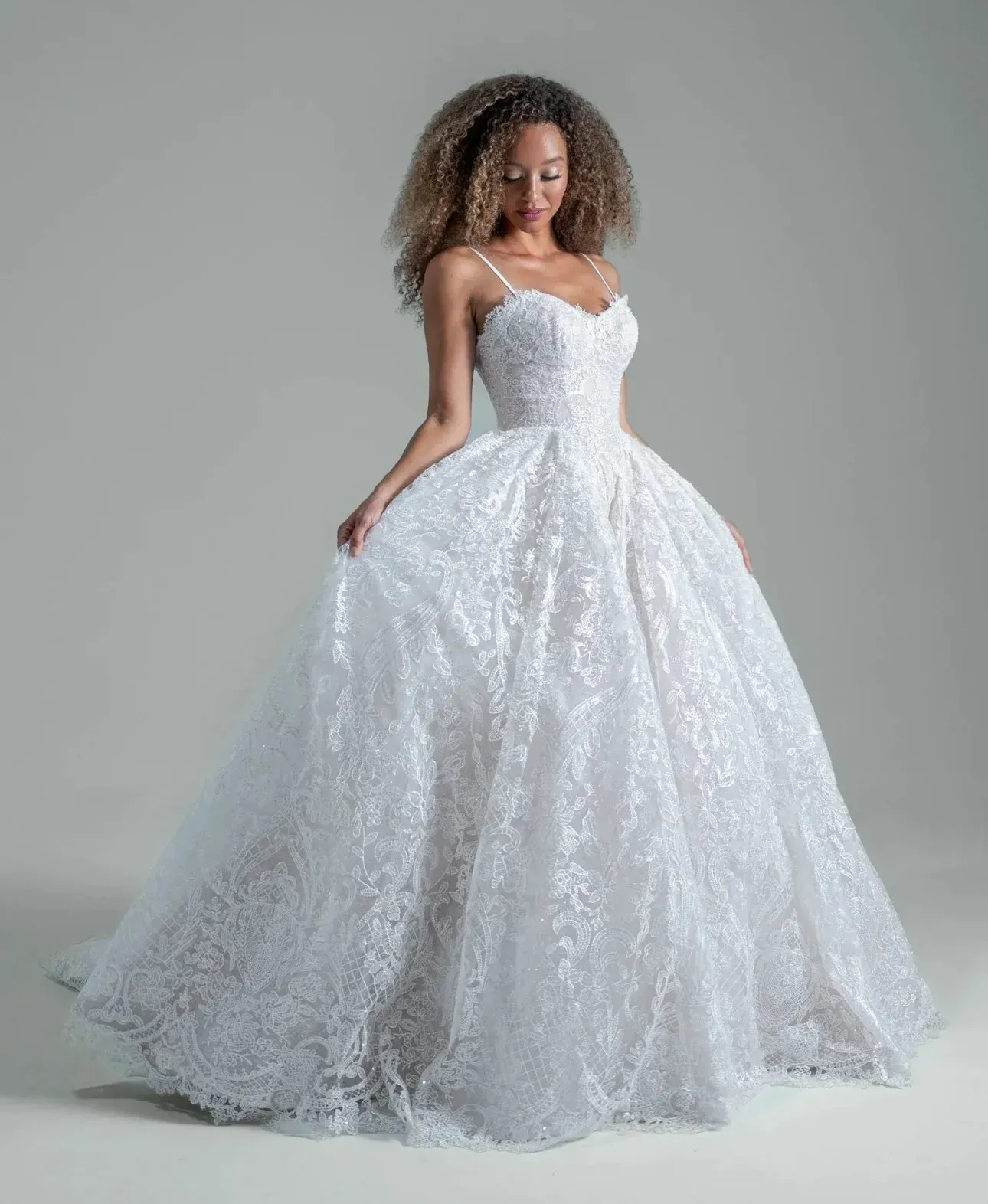 Woman in white lace wedding gown, smiling, holding skirt, standing in studio.