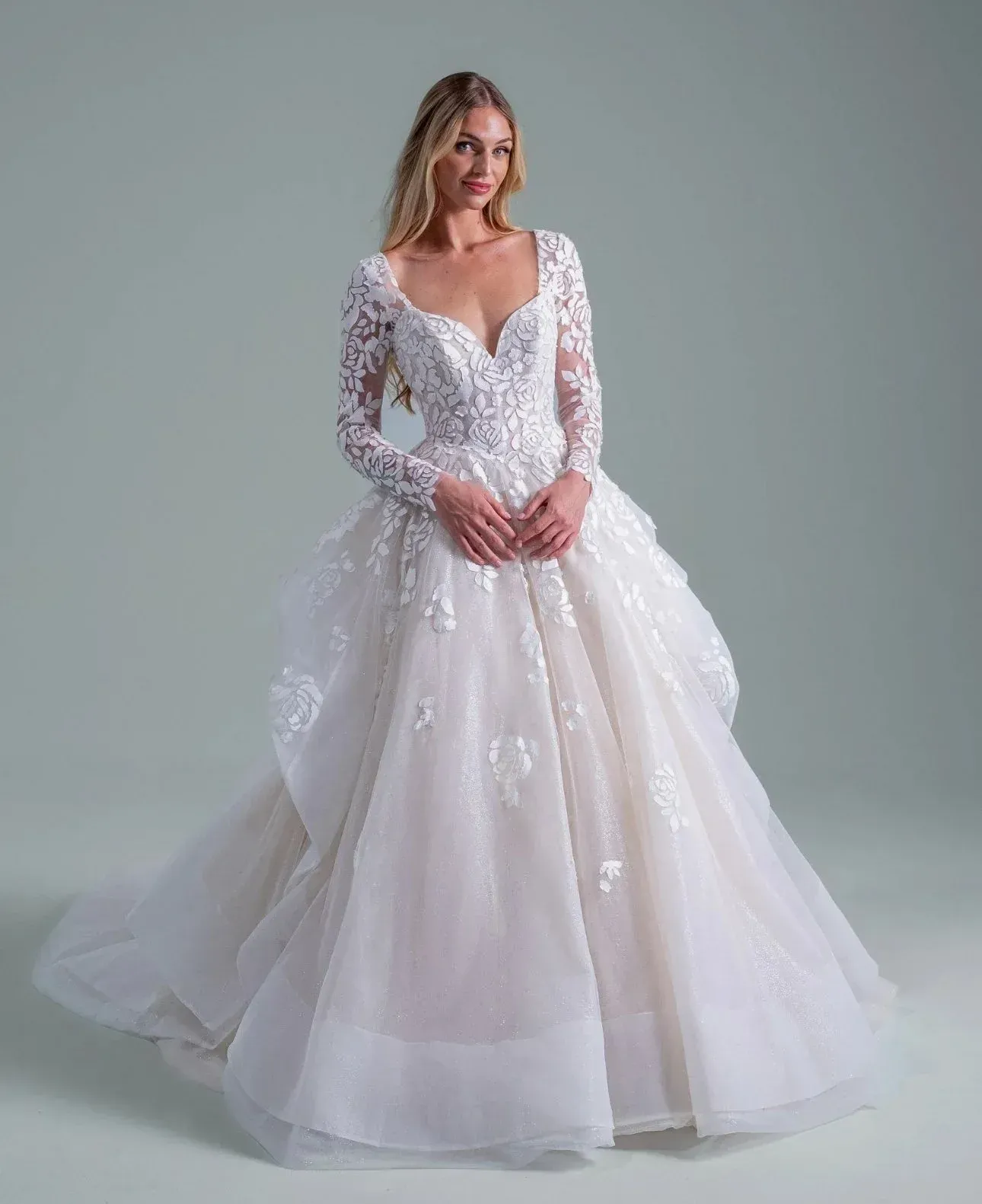 Blonde woman in a white wedding dress with long sleeves, posing in a studio.