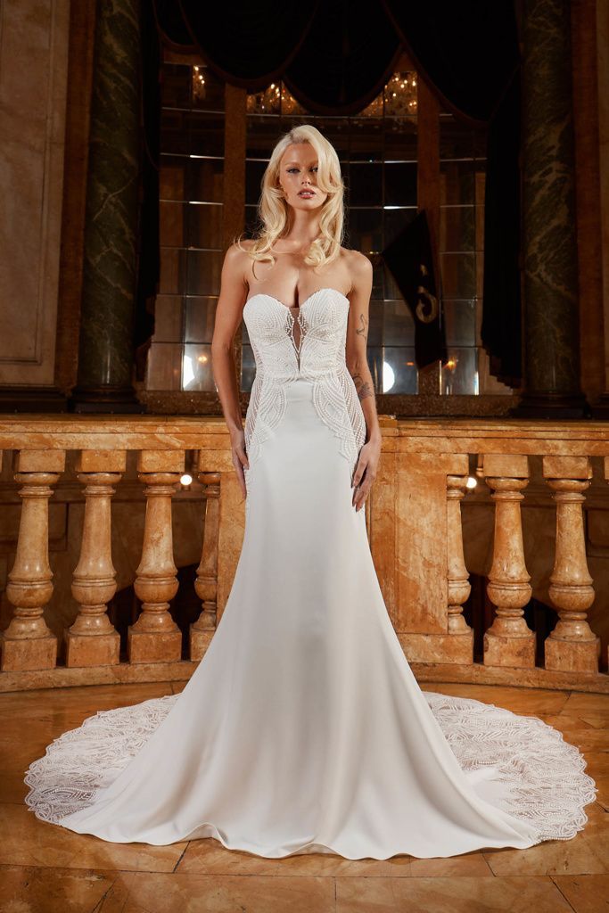 Blonde woman in white wedding dress with high slit, posing in ornate room.
