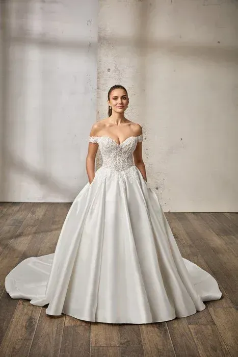 Woman in a white off-the-shoulder wedding dress with a full skirt, posing against a wall.