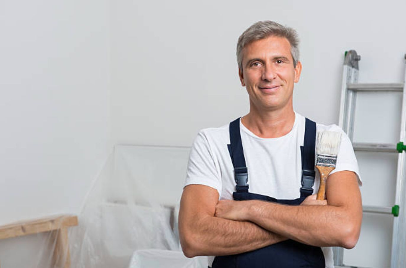 A man is holding a paint brush in a room with his arms crossed.