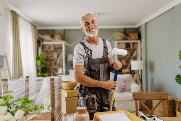 A man is holding a paint roller and a hammer in a living room.