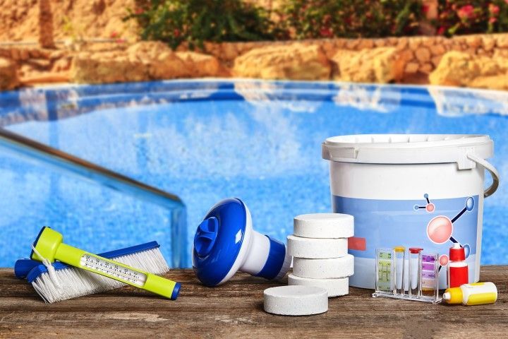 Pool cleaning supplies beside a blue swimming pool: bucket, chemicals, brush, and test tubes
