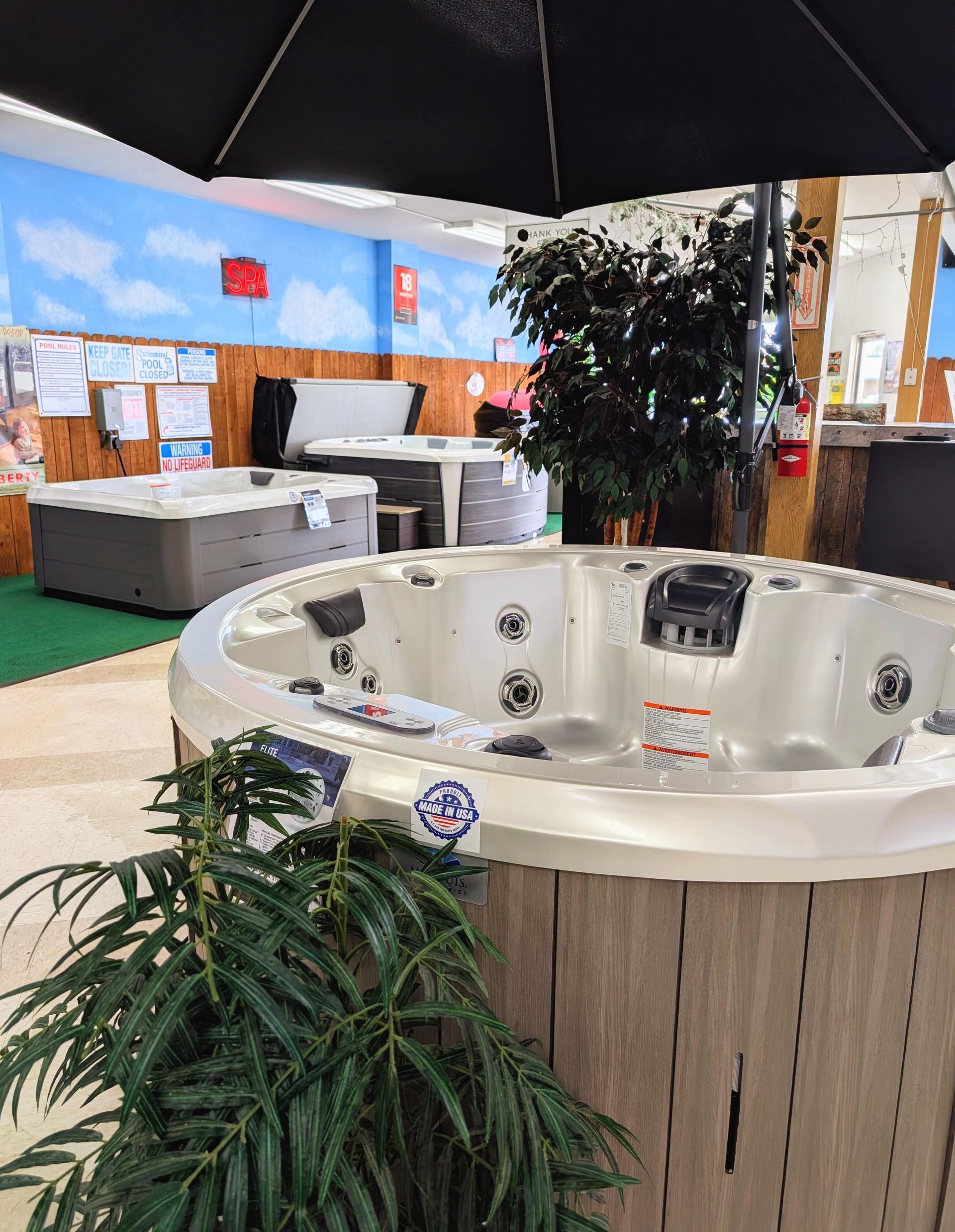 Indoor hot tub showroom displaying multiple spa models including a round white Made in USA hot tub in the foreground with jets and headrests, surrounded by decorative plants and additional spa units on display in the background.