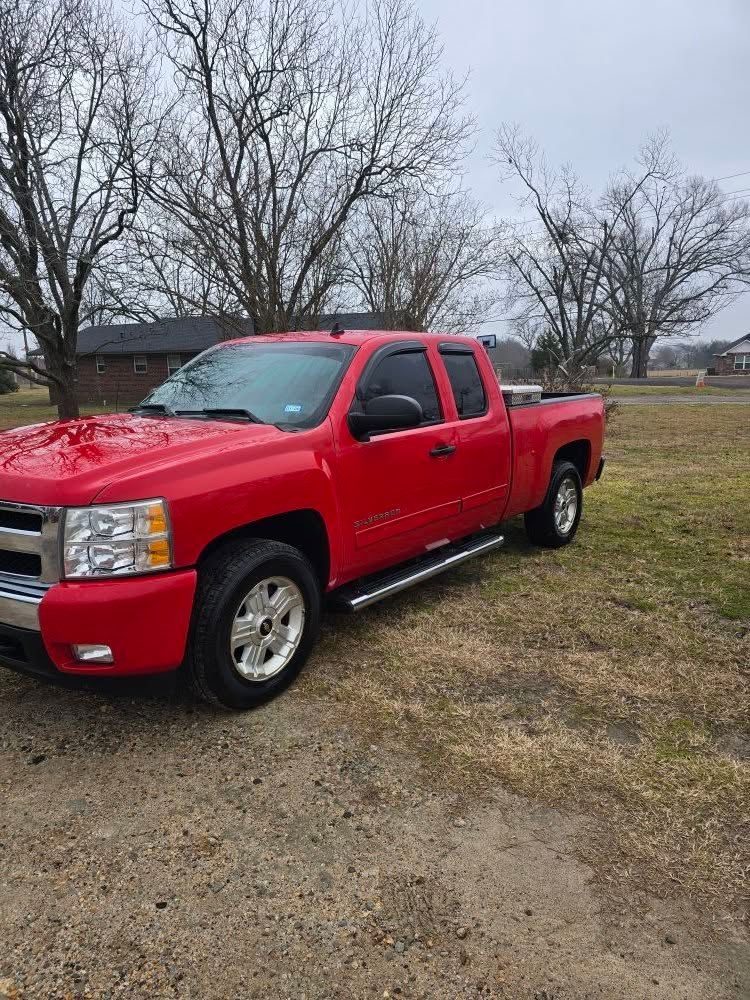 Red Chevrolet pickup truck parked on a grassy lot with bare trees and a house in the background.