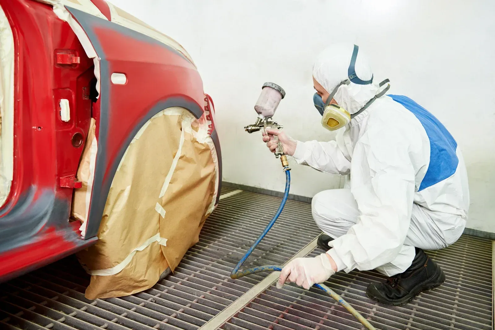 Person in protective gear painting a red car in a body shop.