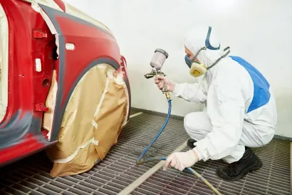 Person in protective gear painting a red car in a body shop.