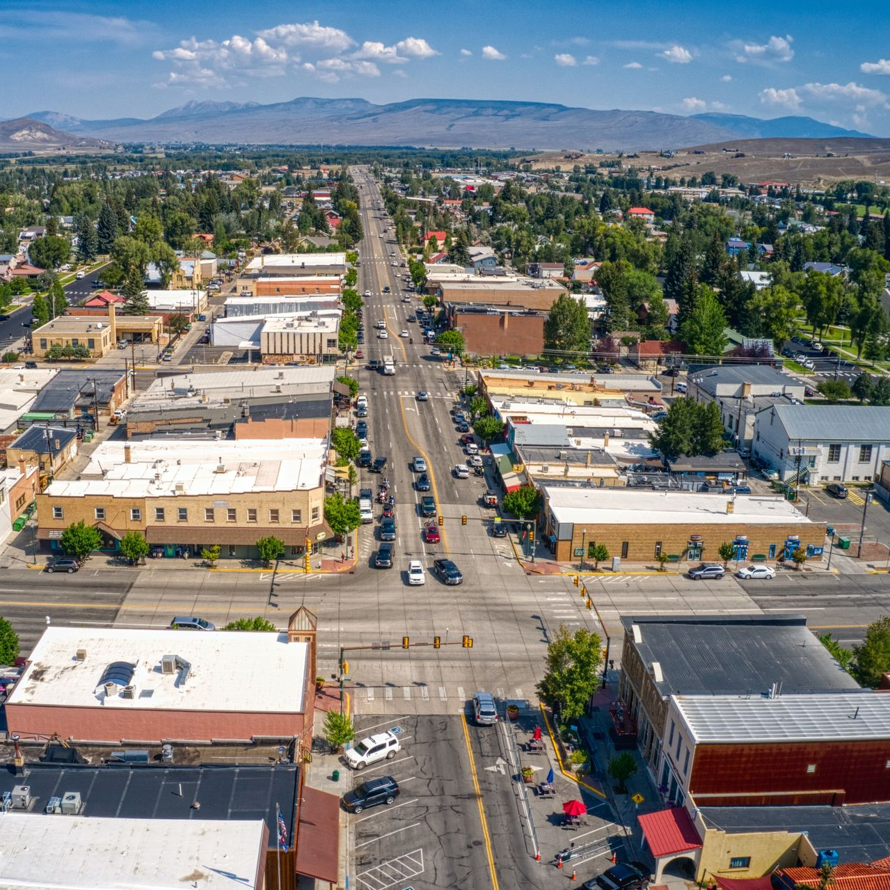 Aerial view of a town with buildings lining a main street, cars driving, and mountains in the background.