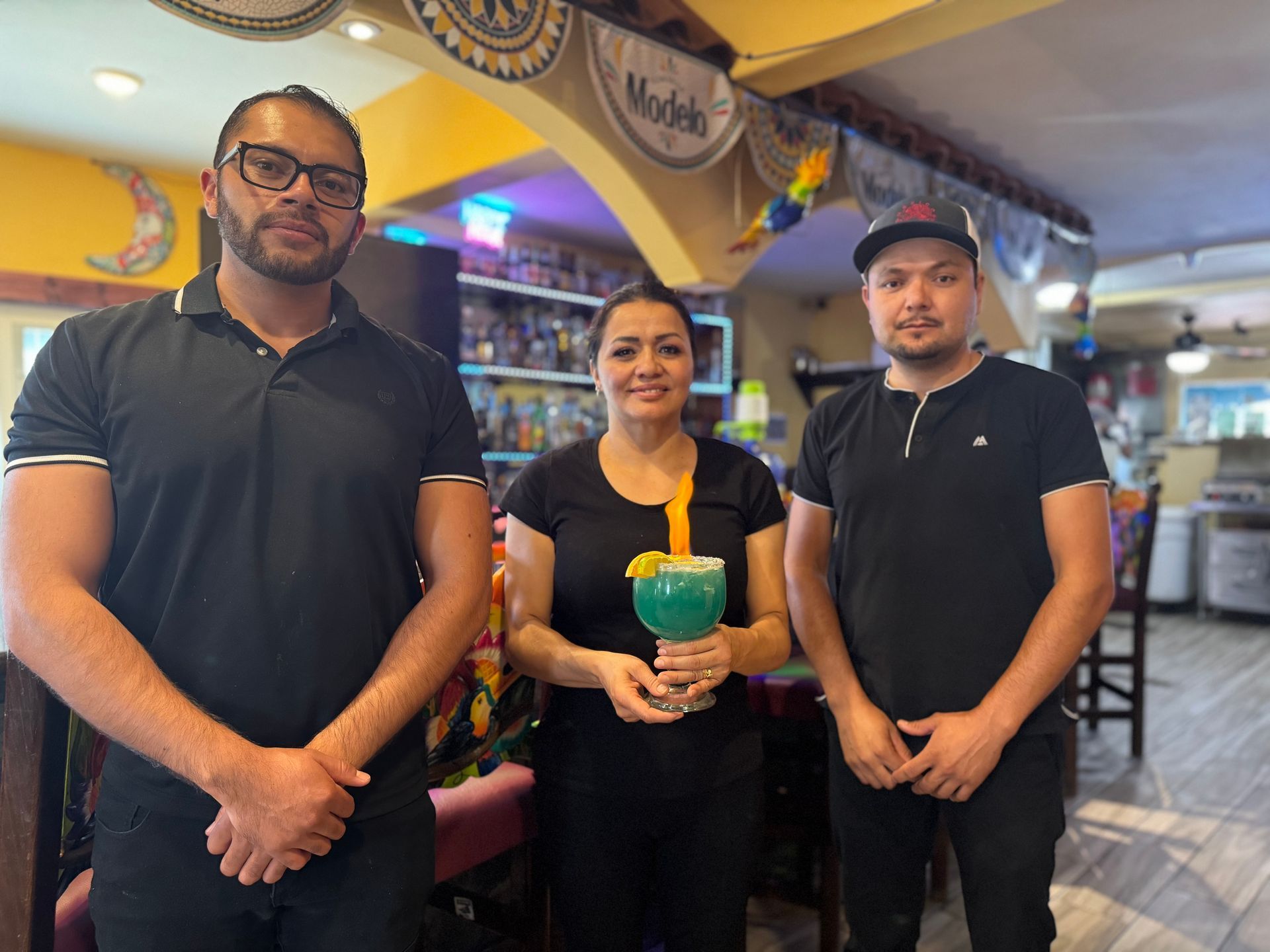 Three people in a restaurant: one holding a drink, two others standing next to her.
