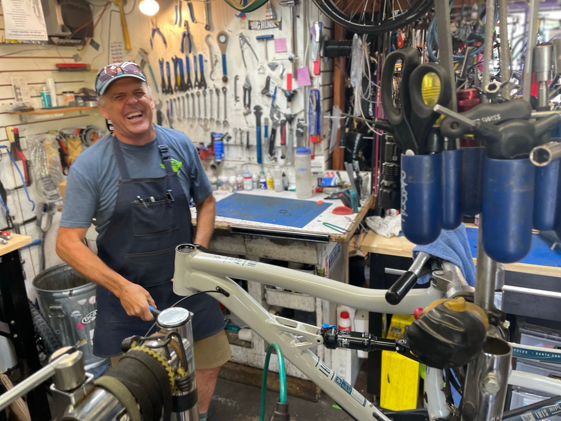 Man in apron smiles while working on a bike frame in a cluttered workshop.