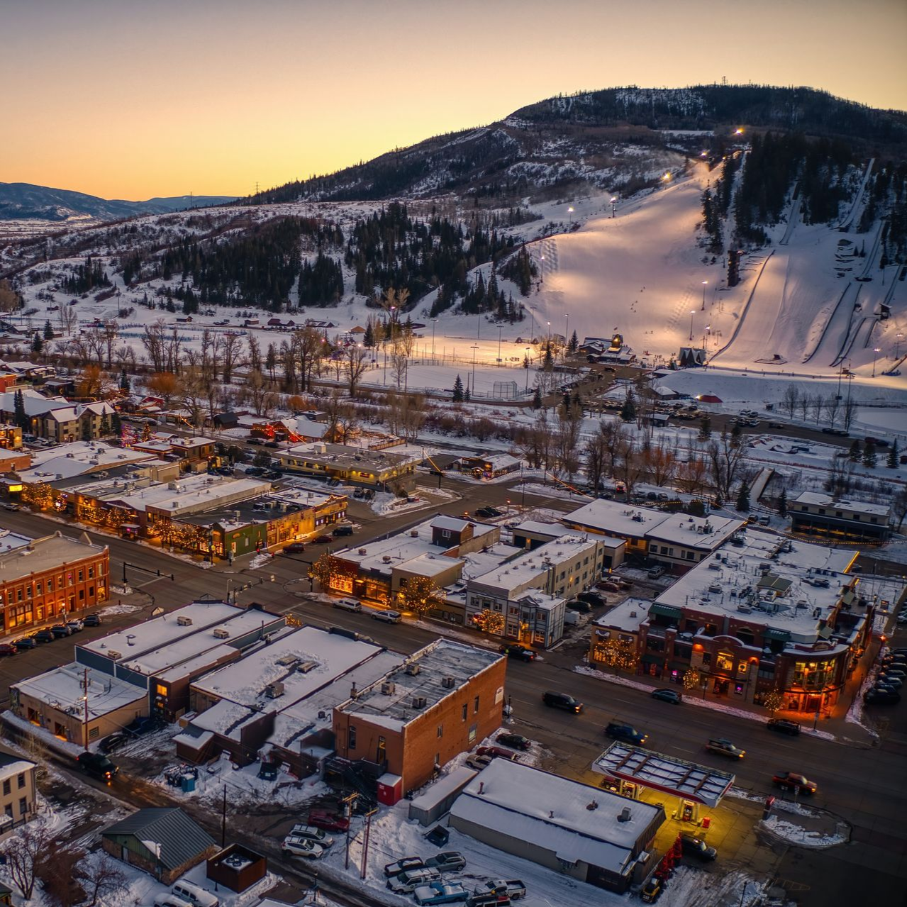 Snowy town at dusk with lit buildings and a ski slope on a mountain in the background.