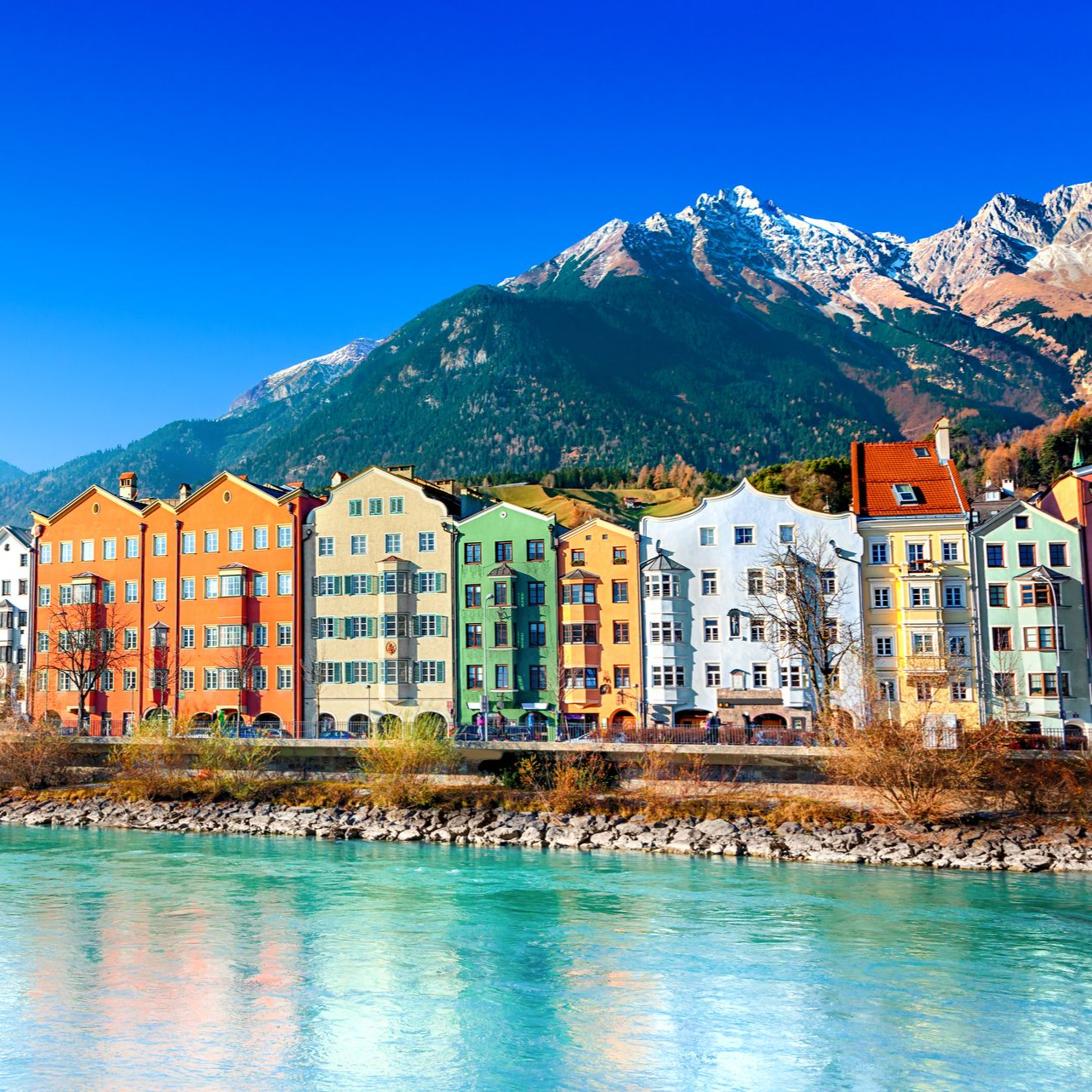 Colorful buildings line a river against a backdrop of mountains under a clear blue sky.