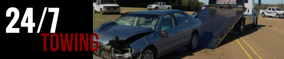 A damaged blue car being towed by a tow truck on a road.