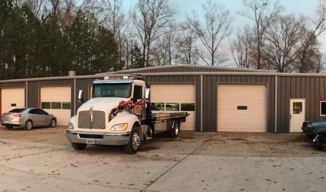 Tow truck in front of a gray metal building with garage doors; a car is parked nearby.