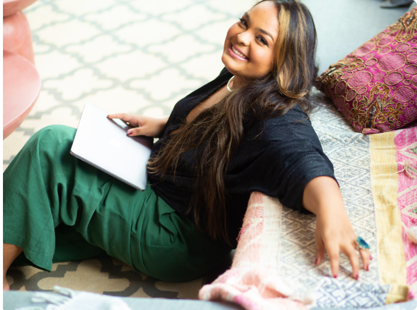 Uma mulher está deitada no chão segurando um laptop e sorrindo.