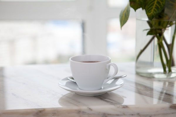 A cup of coffee is sitting on a saucer on a table next to a vase of flowers. - Luther Falls Kitchen & Bath - Champaign, IL