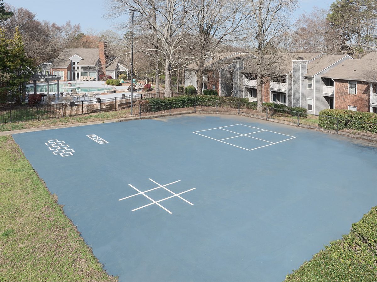 Outdoor blue play area with tic-tac-toe, hopscotch, and four square game markings, surrounded by greenery.