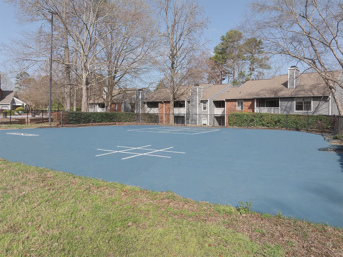 Blue-painted tennis court with patchy white markings, surrounded by grass and apartment buildings.