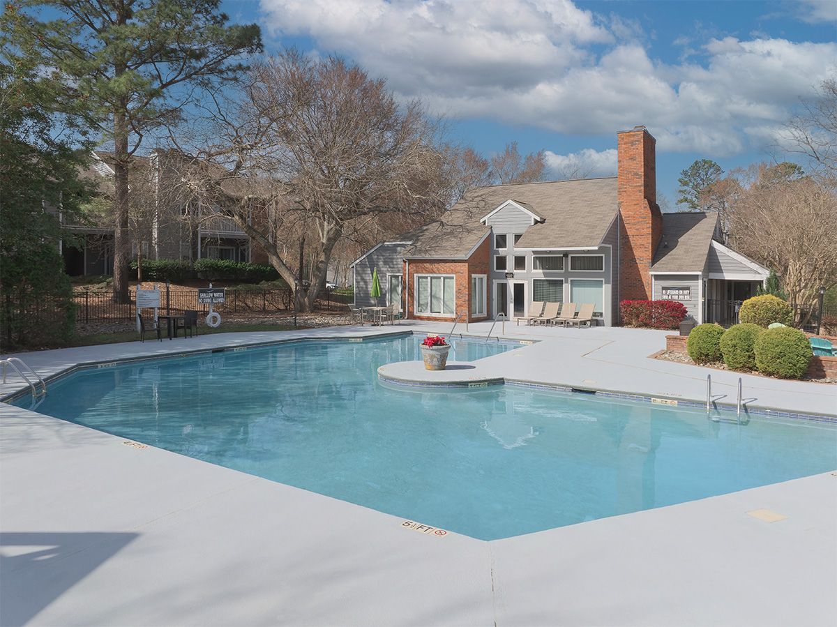 Swimming pool with a house in the background, surrounded by trees. Blue sky with clouds.