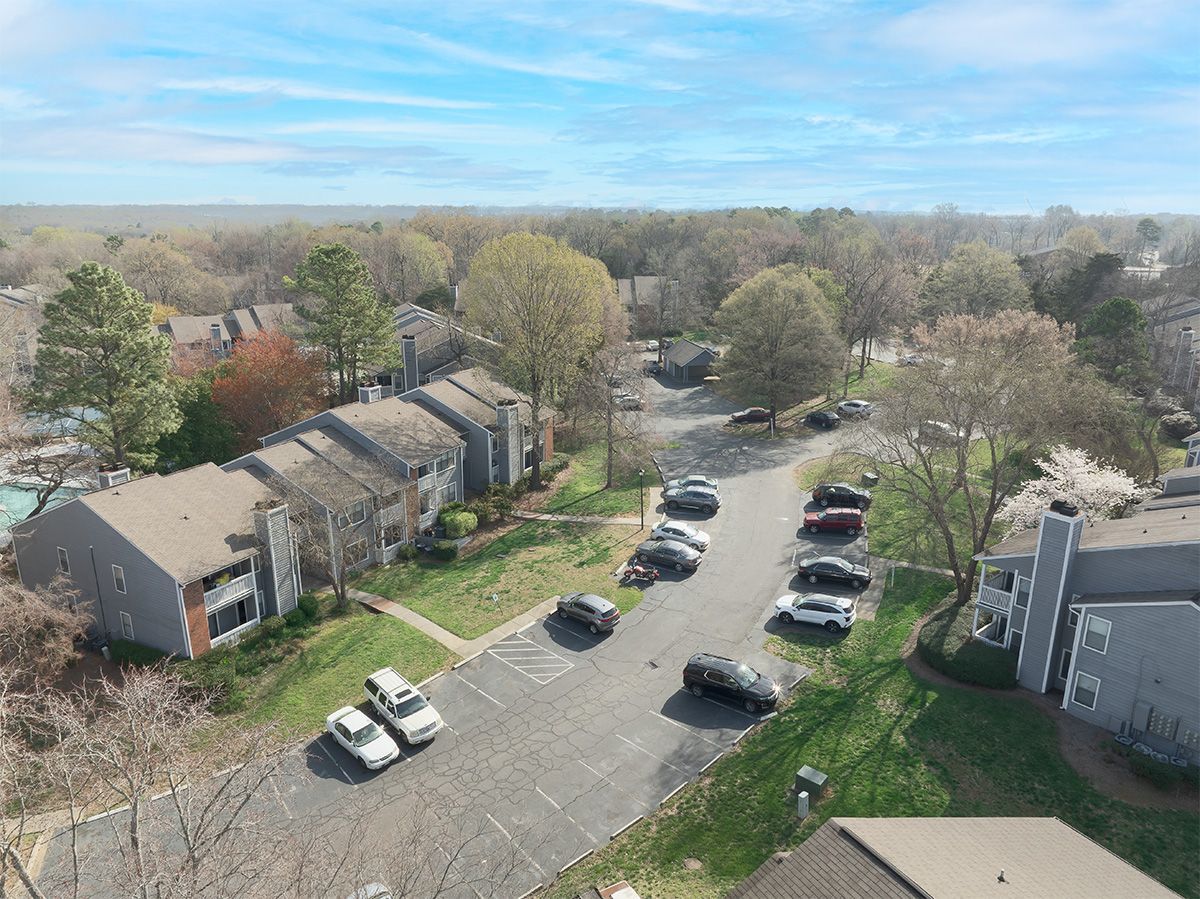 Apartment buildings with parked cars on a paved road, surrounded by trees and grass, under a blue sky.