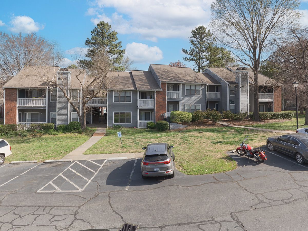 Apartment complex with gray siding, red brick accents, and parked cars on a sunny day.