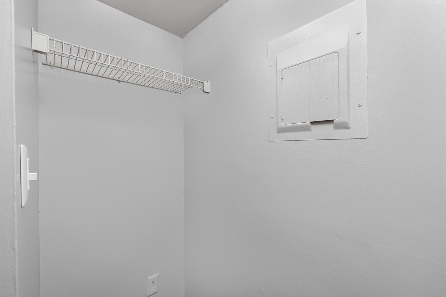 Empty, light-gray painted storage closet. White wire shelf mounted to the wall. White access panel on the right.