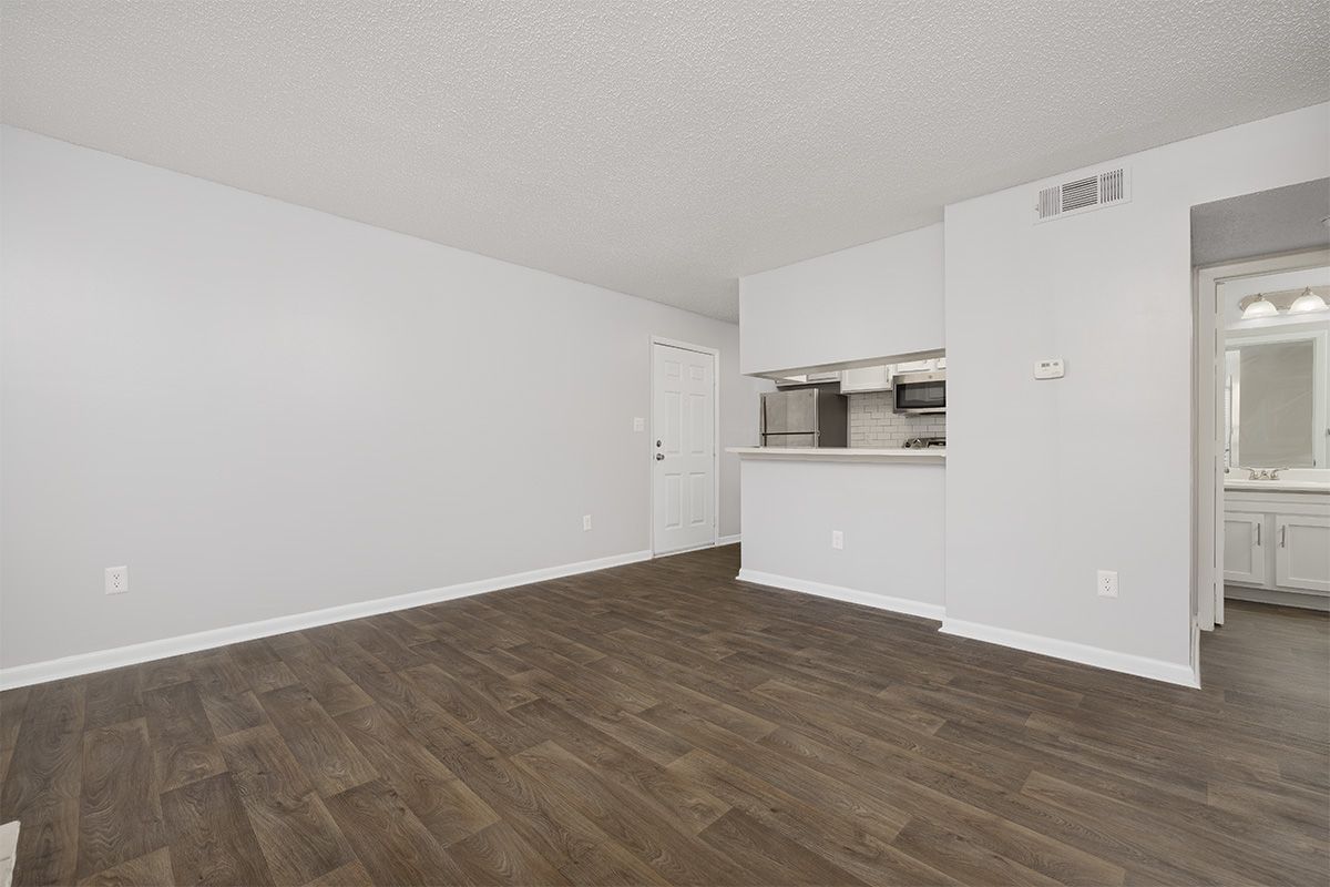 Empty living room with gray walls, dark wood-look floors, and an opening to the kitchen/bathroom.