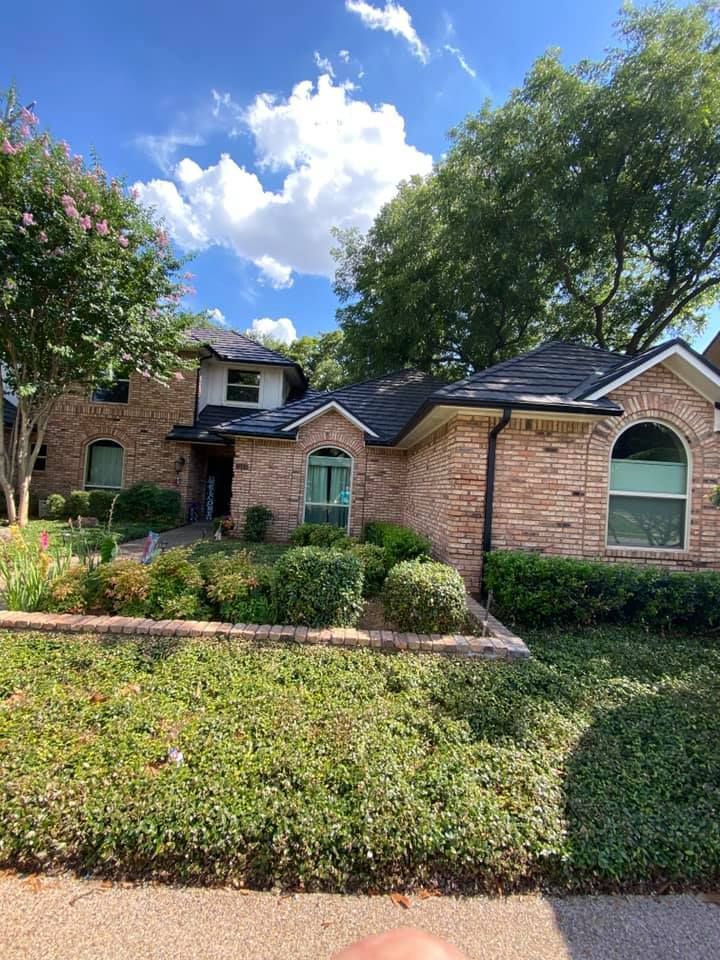 A large brick house with a black roof is surrounded by trees and bushes.
