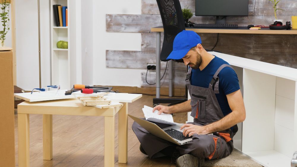 Worker in blue cap sitting on the floor, reading a clipboard in a modern room with tools and a table nearby.