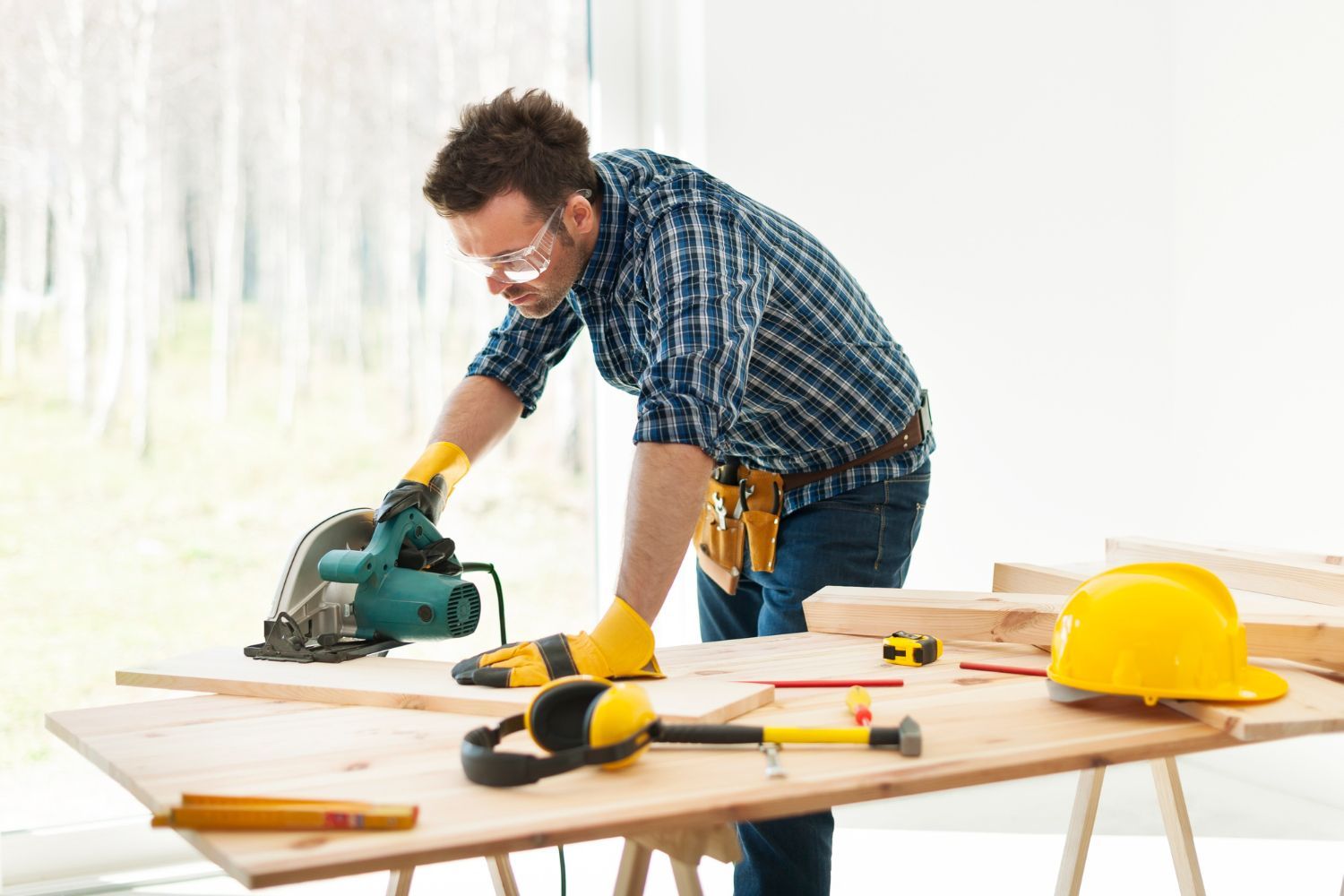 Person using a circular saw at a workbench with tools and a yellow hard hat in a bright workshop