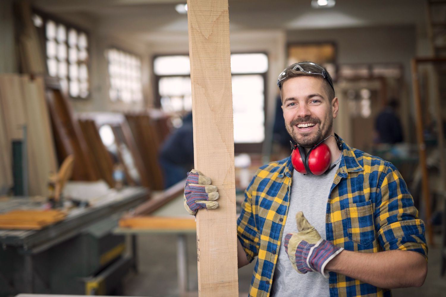 Man in plaid shirt and safety gloves holding a wood plank in a workshop, smiling.