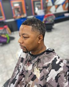 A man with a short, faded haircut and a shaped beard, wearing a camouflage barber cape inside a brightly lit salon.
