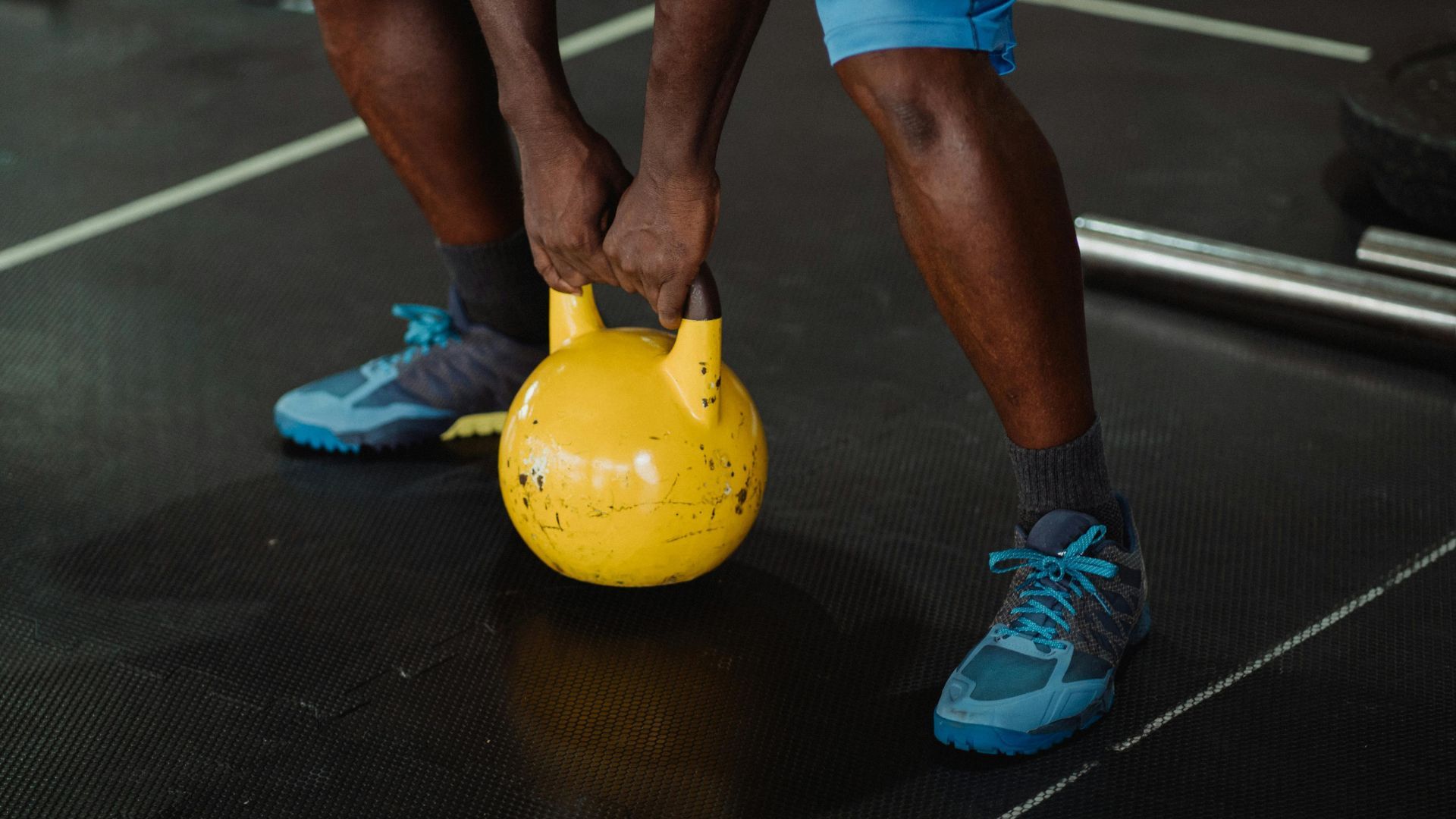 A person in blue athletic shoes stands on a gym floor, gripping the handle of a yellow kettlebell.
