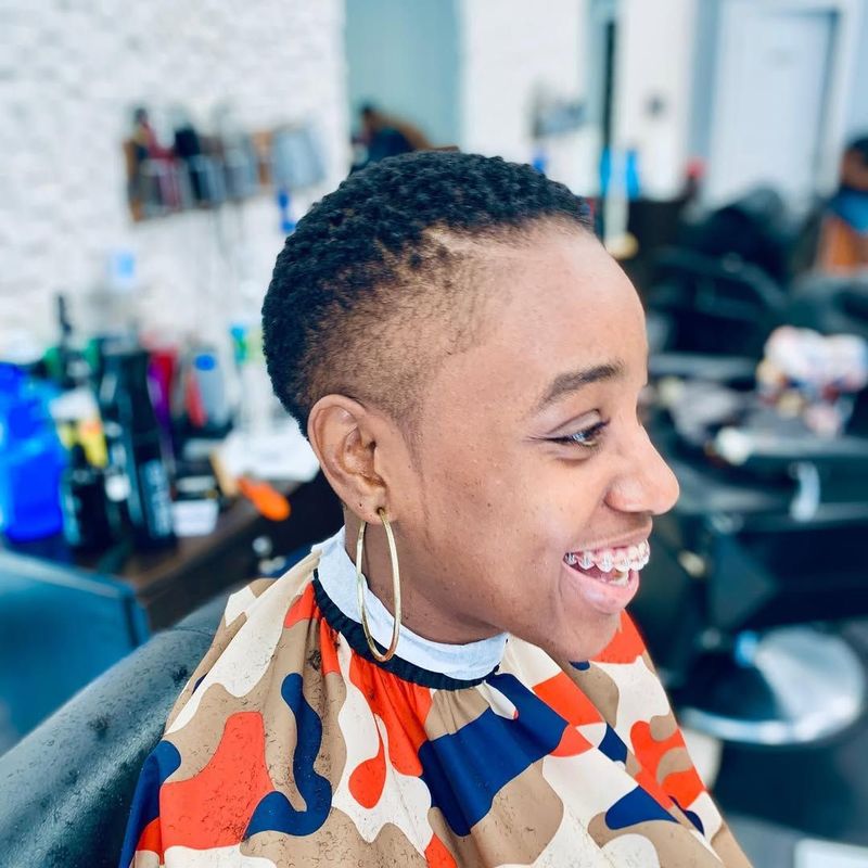 A person wearing a camo cape smiles in a barbershop with a short, tapered haircut.