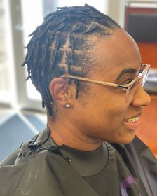 A person with short, parted locs wearing glasses, smiling in a salon cape.