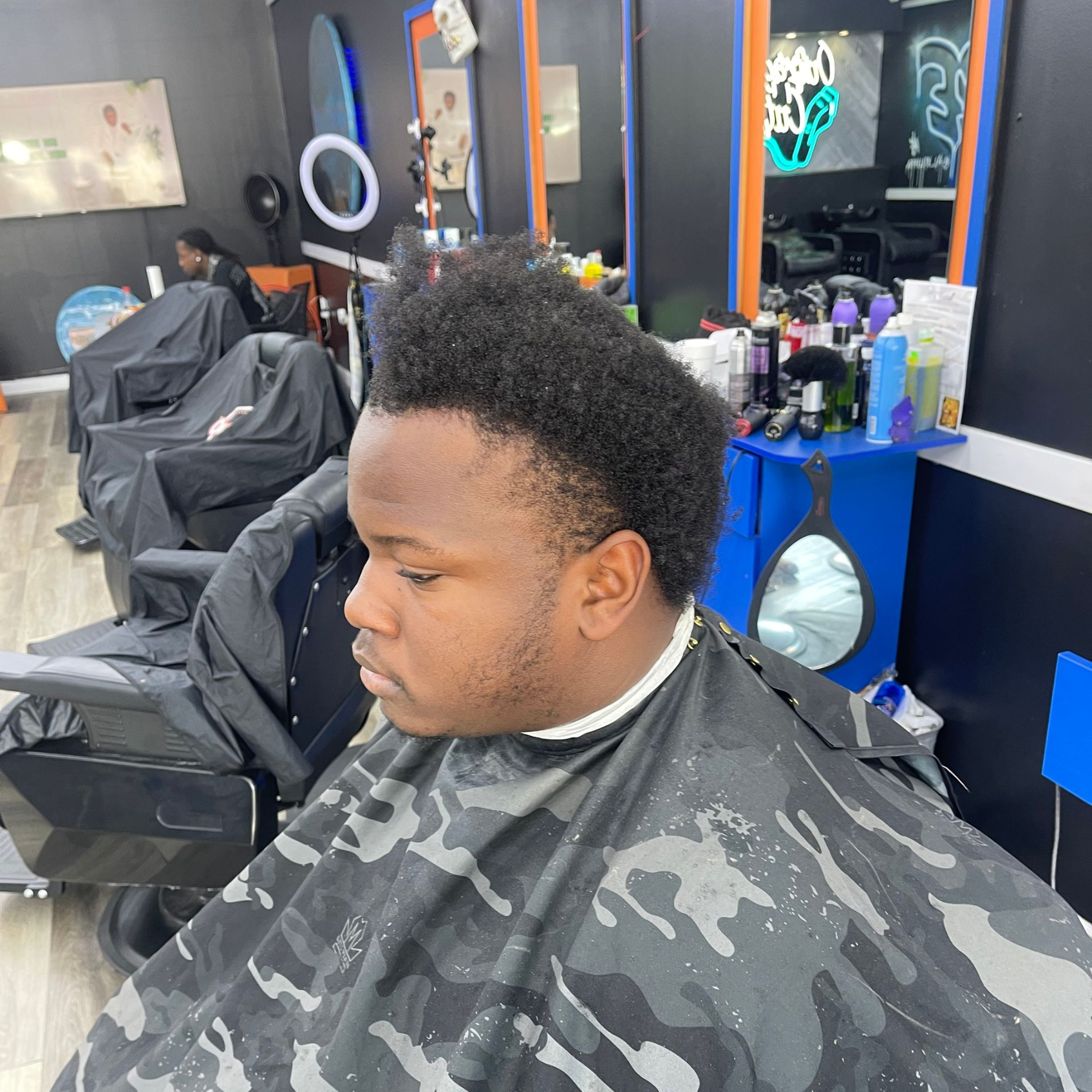Client seated in a barbershop chair with a dark patterned cape, surrounded by mirrors and grooming products.