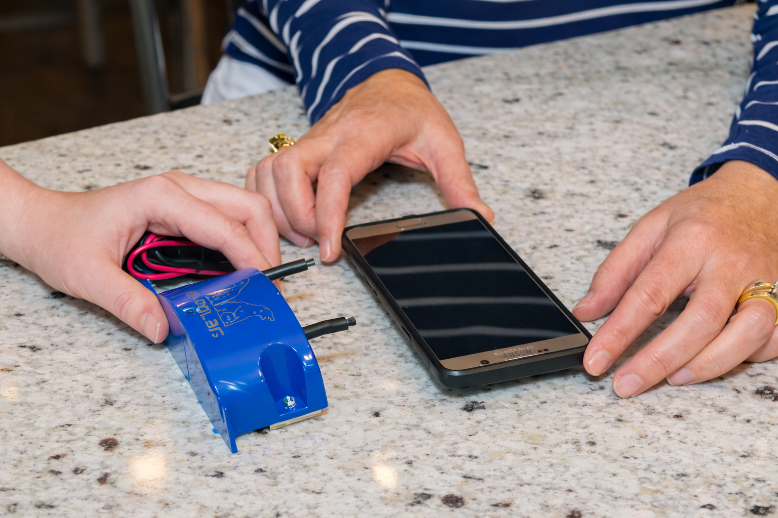 Two hands placing a blue device near a smartphone on a speckled countertop.