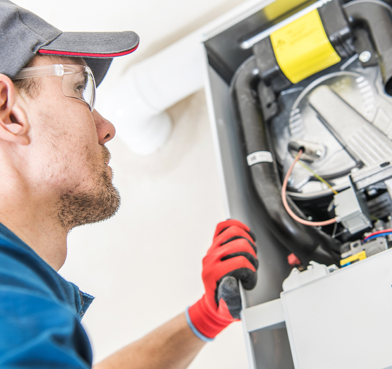 HVAC technician inspecting furnace, wearing safety glasses and gloves, using a tool.