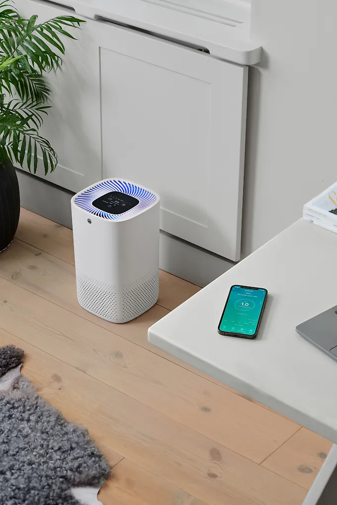 White air purifier next to a desk with a smartphone displaying an app; a plant in the background.