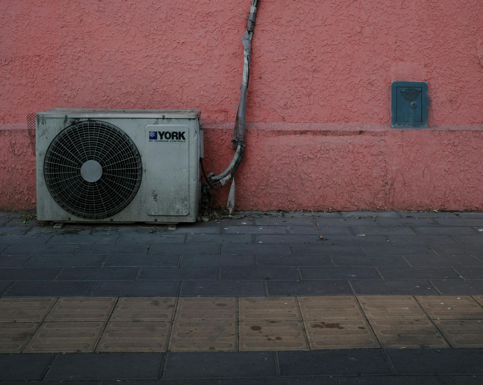 Air conditioning unit mounted on a textured, pink wall above a tiled sidewalk.