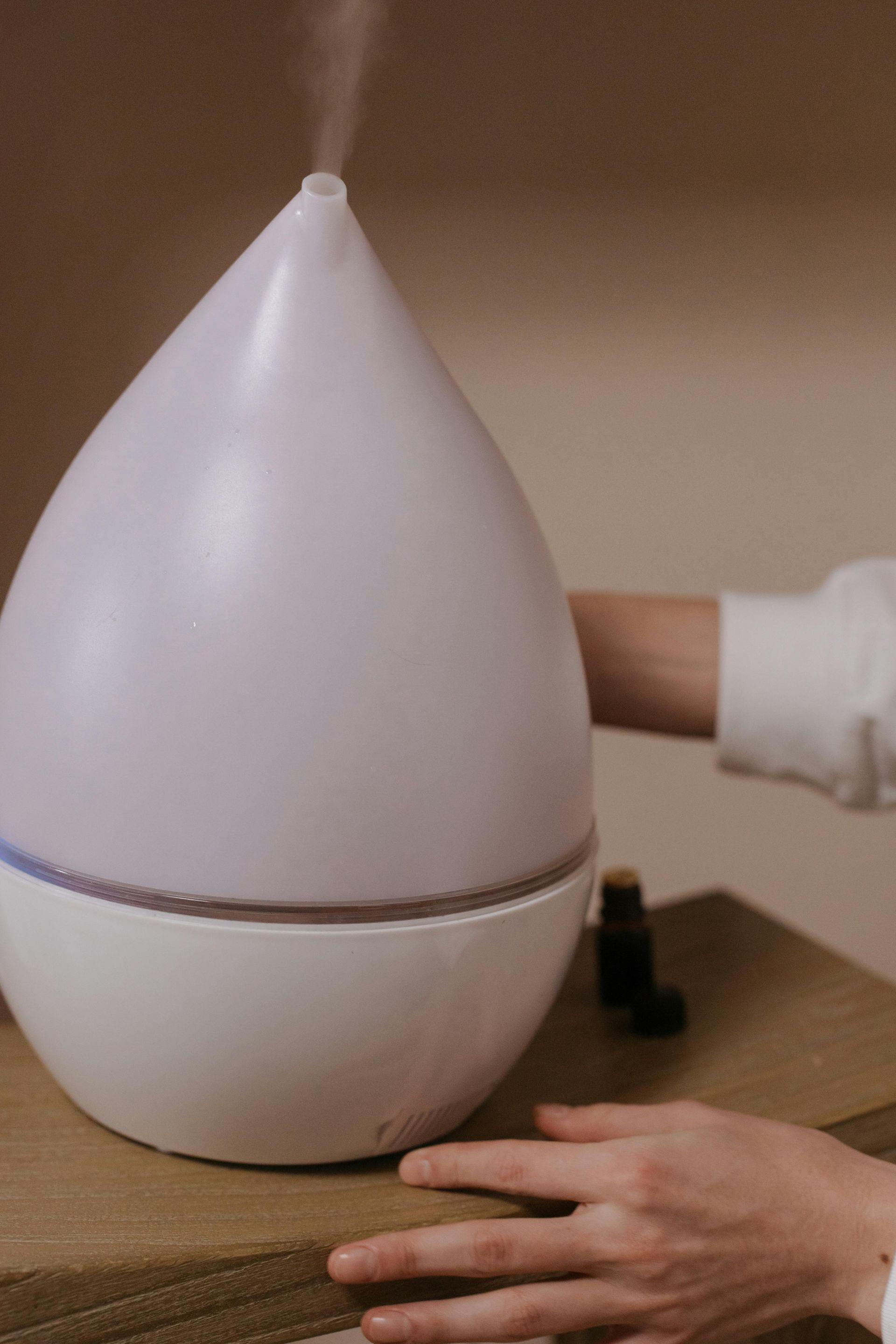 White and purple teardrop-shaped essential oil diffuser with steam. A hand reaches toward it; small bottle visible.