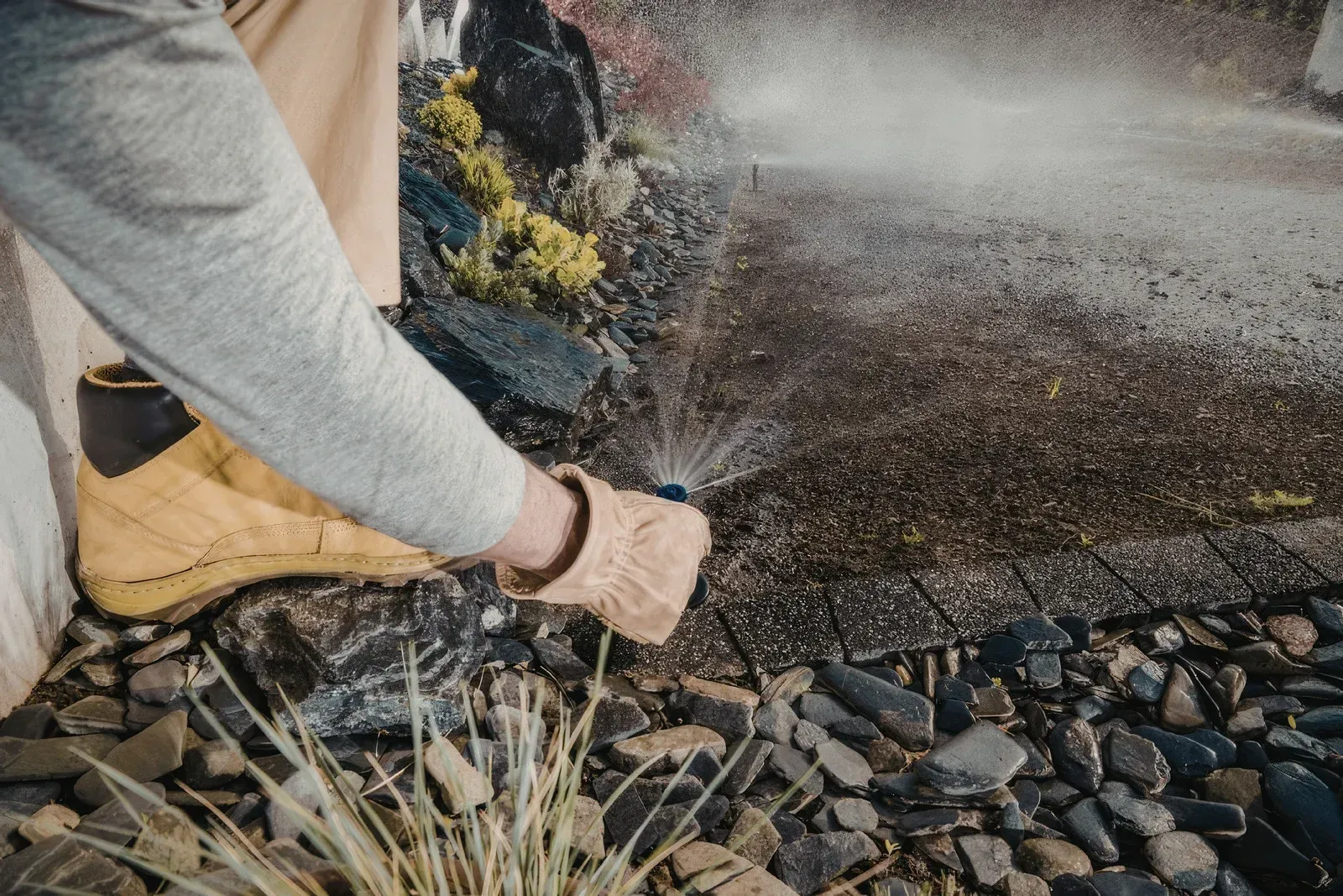 A person wearing work gloves and tan boots sprays water onto a gravel garden bed near a paved surface.